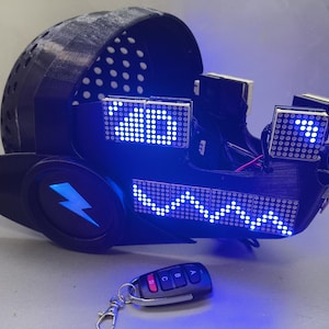 May include: A black, futuristic helmet with blue LED lights and a lightning bolt design. The helmet has multiple LED displays and a remote control. The overall design suggests a cyberpunk or sci-fi theme.