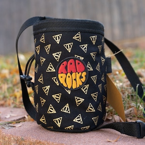 May include: Black chalk bag featuring a gold triangle pattern and a circular patch with the text "RAD ROCKS". It has a black strap and drawstring closure. A brown tag is attached.