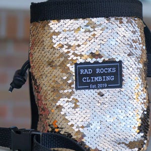 Puede incluir: Una bolsa de tiza de escalada con un exterior de lentejuelas doradas. La bolsa tiene un cierre de cordón negro, una correa negra con hebilla y un borde negro. Un parche rectangular en la parte delantera dice "RAD ROCKS CLIMBING Est. 2019".