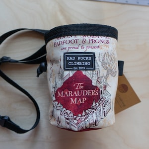 May include: A cream-colored chalk bag with a black drawstring closure and strap. The bag features a map design with the text "The Marauder's Map" and "Rad Rocks Climbing Est. 2019". A small tag hangs from the side.