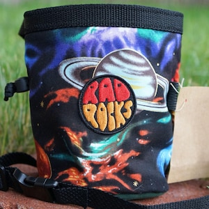 Könnte beinhalten: Schwarzer Chalkbag mit einem lebendigen Weltraumdesign mit Planeten und Nebeln. Der Beutel hat einen schwarzen Kordelzugverschluss und einen verstellbaren schwarzen Riemen. Ein kreisförmiger Aufnäher mit der Aufschrift "RAD ROCKS" ist auf der Vorderseite aufgenäht.