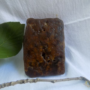 Puede incluir: Una pastilla de jabón rectangular de color marrón oscuro con una superficie texturizada y agujeros irregulares. El jabón está sobre una tela blanca, con una hoja verde y una pequeña rama en primer plano.