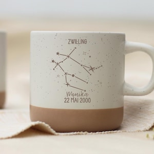 May include: A speckled white ceramic mug with a brown base. The mug features a constellation design with the word "ZWILLING" and the name "Monika" with the date "22 MAI 2000" printed on it. The mug has a curved handle.