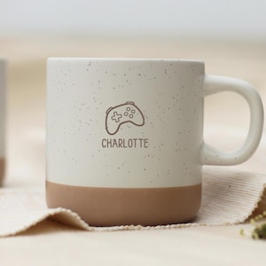 Könnte beinhalten: Ein Keramikbecher mit gesprenkeltem cremefarbenem Oberteil und einem einfarbigen hellbraunen Boden. Auf der Vorderseite befindet sich eine braune Strichzeichnung eines Gamecontrollers über dem Namen CHARLOTTE in brauner Schrift.