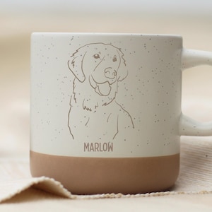 Könnte beinhalten: Eine Keramiktasse mit braunem Boden und einem gesprenkelten cremefarbenen oberen Bereich. Die Tasse zeigt eine Strichzeichnung eines Hundegesichts und den Namen "MARLOW". Die Tasse hat einen gebogenen Henkel und steht auf einer neutralen Oberfläche.