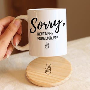 Könnte beinhalten: Weiße Keramik-Tasse mit dem Text "Sorry, NICHT MEINE ENTGELTGRUPPE." in schwarzer Schrift und einem Peace-Zeichen. Ein Holzuntersetzer mit passendem Peace-Zeichen ist ebenfalls zu sehen.