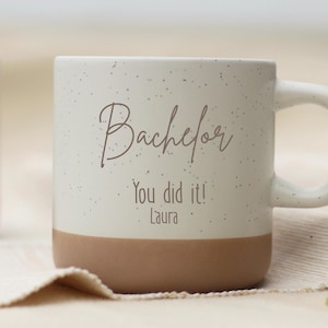 Könnte beinhalten: Eine gesprenkelte weiße Keramik-Tasse mit braunem Boden und Henkel. Auf der Tasse stehen die Worte "Bachelor", "You did it!" und "Laura". Die Tasse steht auf einem beigen Tuch.