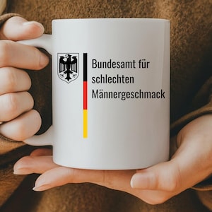 Könnte beinhalten: Weiße Keramiktasse mit einem schwarz-gelben deutschen Flaggen-Design und dem aufgedruckten Text "Bundesamt für schlechten Männergeschmack".