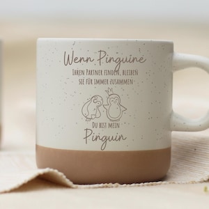 Könnte beinhalten: Eine weiße Keramik-Tasse mit gesprenkelter Oberfläche und braunem Boden. Die Tasse zeigt eine Pinguin-Illustration und deutschen Text. Der Griff ist weiß. Die Tasse steht auf einem beigen Tuch.