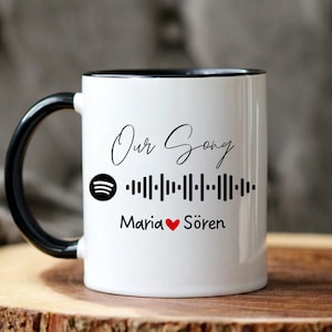 Könnte beinhalten: Weißer Keramikbecher mit schwarzem Henkel und Rand. Der Becher zeigt den Text "Our Song" in Schreibschrift, ein Spotify-Logo, einen Barcode und die Namen "Maria" und "Sören" mit einem Herzsymbol. Der Becher steht auf einer Holzoberfläche.