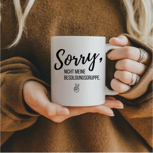 May include: White ceramic mug with the text "Sorry, NICHT MEINE BESOLDUNGSGRUPPE." in black script. A peace sign is below the text. The mug is held by a person wearing a brown sweater.