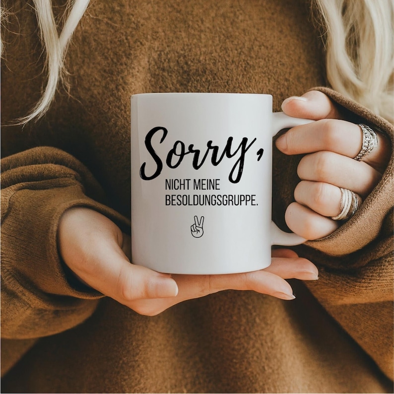 May include: White ceramic mug with the text "Sorry, NICHT MEINE BESOLDUNGSGRUPPE." in black script. A peace sign is below the text. The mug is held by a person wearing a brown sweater.