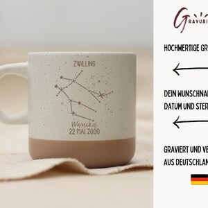 May include: A speckled ceramic mug with a white handle and a tan base. The mug features an engraved Gemini constellation design, the name "Monika", and the date "22 MAI 2000".