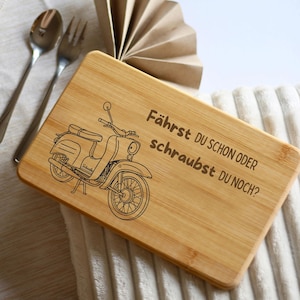 May include: A rectangular bamboo cutting board with a black line drawing of a vintage scooter and the German text "Fährst du schon oder schraubst du noch?" A spoon and fork are visible in the upper left corner.