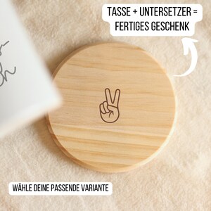 May include: A round, light-colored wooden coaster with a peace sign graphic. The coaster has a natural wood grain pattern and is shown on a textured, off-white surface. Text in German reads "TASSE + UNTERSETZER = FERTIGES GESCHENK" and "W&Auml;HLE DEINE PASSENDE VARIANTE".