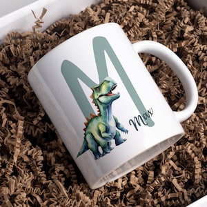 Könnte beinhalten: Weiße Keramiktasse mit einem grünen Buchstaben M und einer Aquarell-Illustration eines grünen Dinosauriers. Die Tasse trägt den Namen Max.
