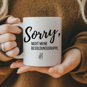 Könnte beinhalten: Weiße Keramik-Tasse mit den Worten "Sorry, Nicht meine Besoldungsgruppe" in schwarzer Schrift. Darunter ein Friedenszeichen. Die Tasse wird von einer Person gehalten, die einen braunen Pullover trägt.