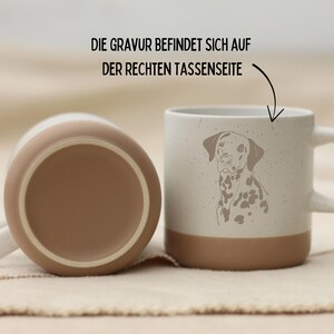 May include: Two ceramic mugs. One is upside down, showing a brown base. The other mug is upright, featuring a Dalmatian dog illustration. The mug has a white handle and a brown base. The text on the image says, "The engraving is on the right side of the cup."