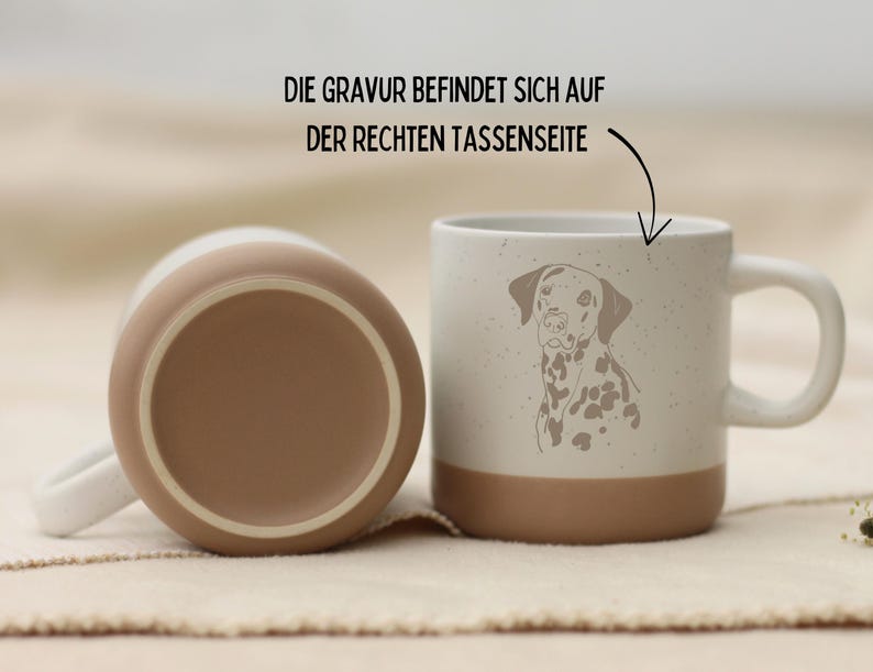 May include: Two ceramic mugs. One is upside down, showing a brown base. The other mug is upright, featuring a Dalmatian dog illustration. The mug has a white handle and a brown base. The text on the image says, "The engraving is on the right side of the cup."