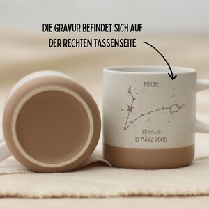 May include: Two ceramic mugs, one upright and one inverted, with a white and tan color scheme. The upright mug features a constellation design with the word "Fische" and the name "Maria" with a date. The text "DIE GRAVUR BEFINDET SICH AUF DER RECHTEN TASSENSEITE" is also visible.