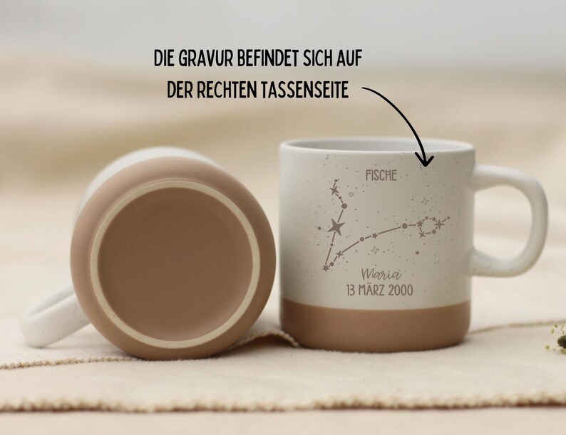 May include: Two ceramic mugs, one upright and one inverted, with a white and tan color scheme. The upright mug features a constellation design with the word "Fische" and the name "Maria" with a date. The text "DIE GRAVUR BEFINDET SICH AUF DER RECHTEN TASSENSEITE" is also visible.