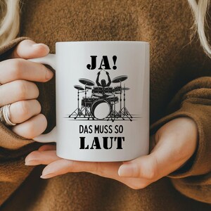 Könnte beinhalten: Weiße Keramiktasse mit einer schwarzen Silhouette eines Schlagzeugers, der ein Schlagzeug spielt. Die Tasse trägt den Text "JA! DAS MUSS SO LAUT".