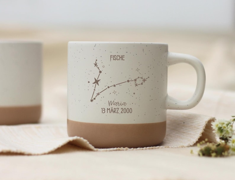 May include: A white ceramic mug with a speckled finish and a brown base. The mug features a constellation design with the word "FISCHE" and the name "Maria" with the date "13 M&auml;rz 2000".