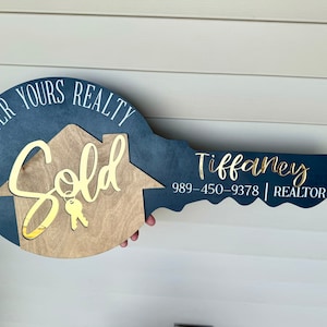 May include: A blue and wood key-shaped sign with the words "Forever Yours Realty" and "Sold" in white lettering. The sign also includes the name "Tiffany" and the phone number 989-450-9378, along with the word "REALTOR" in white lettering.