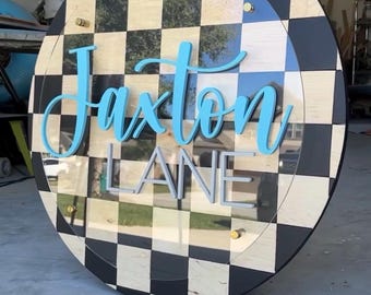 May include: A round wooden sign with a black and white checkered border. The sign features the name "Jaxton" in light blue cursive lettering above the word "LANE" in gray block letters, all set against a clear acrylic background.
