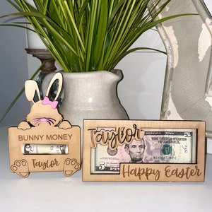 May include: Two wooden money holders with the name "Taylor" engraved on them. One holder is shaped like a bunny with a pink bow and the text "Bunny Money". The other holder has the text "Happy Easter".