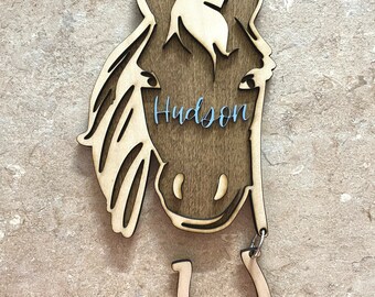 May include: Wooden horse head cutout with the name "Hudson" engraved on it. A wooden horseshoe with the text "1 Month" is attached to the bottom of the horse head.