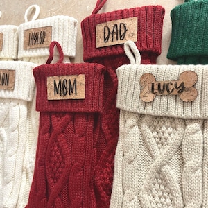 May include: Five knitted Christmas stockings with personalized cork name tags. The stockings are white, red, and green. The names on the tags are Tatum, Taylor, Mom, Dad, and Lucy.