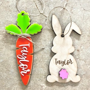 May include: Two Easter-themed wooden ornaments. One is a carrot with green leaves and the name "Taylor" in white. The other is a bunny with a pink tail and the name "Taylor". Both have twine loops for hanging.
