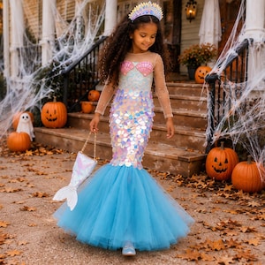 May include: A mermaid costume featuring a bodice with pink shell accents, long sheer sleeves, and a skirt covered in iridescent sequins that transition into a full, light blue tulle hem. The outfit includes a matching crown and a small fish-shaped bag.