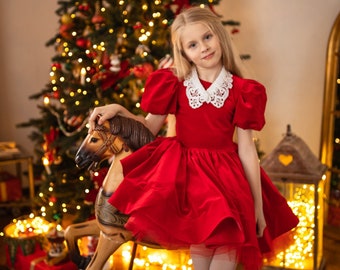 May include: A young girl in a red dress with a white lace collar sits on a rocking horse in front of a decorated Christmas tree.