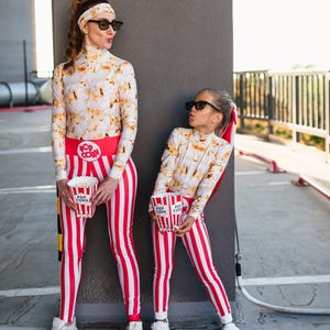 May include: Two people wearing matching popcorn-themed outfits. The outfits include a long-sleeved top with a popcorn print, red and white striped leggings, and a red belt with "Pop Corn" text. Each person holds a popcorn container.