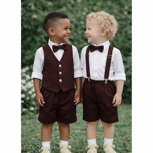 May include: Two children wearing white long-sleeved button-down shirts with dark brown shorts. One child wears a dark brown vest with three gold buttons and a matching bow tie. The other child wears dark brown suspenders and a matching bow tie.
