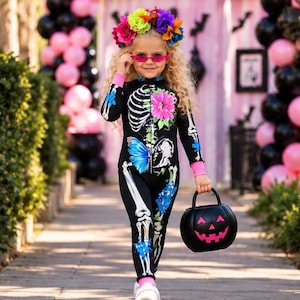 May include: A black long-sleeve jumpsuit featuring a white skeleton print, a vibrant pink flower, and a blue butterfly graphic. The outfit includes pink wrist cuffs and is paired with a black pumpkin-shaped bucket and a colorful floral crown.