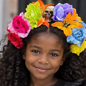 May include: A floral crown featuring a central skull ornament surrounded by artificial flowers in vibrant shades of red, pink, lime green, orange, purple, blue, and yellow. The arrangement consists of various rose and lily-like blossoms.