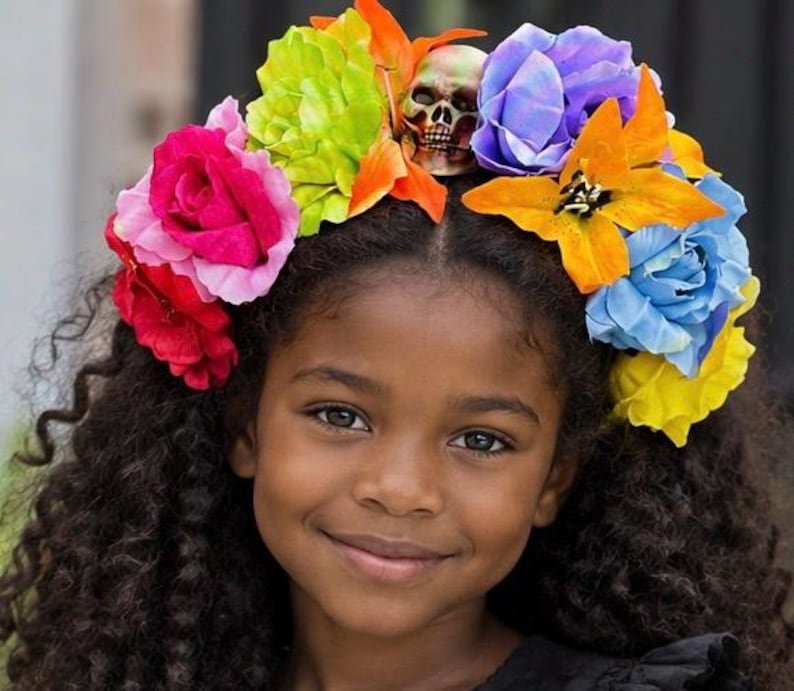 May include: A floral crown featuring a central skull ornament surrounded by artificial flowers in vibrant shades of red, pink, lime green, orange, purple, blue, and yellow. The arrangement consists of various rose and lily-like blossoms.