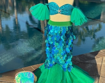 May include: A mermaid costume with a teal sequined top and a matching skirt. The skirt is adorned with blue and teal sequins and a green tulle tail. A small, iridescent purse with a gold chain sits nearby. A pearl necklace is around the mannequin's neck.