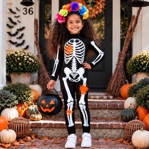 May include: A black long-sleeved jumpsuit featuring a white skeleton print with orange paint splatter accents, paired with a colorful floral crown. The jumpsuit has a fitted silhouette and is worn with white sneakers while holding a plastic pumpkin bucket.