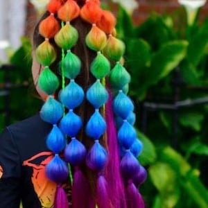 May include: A hairstyle featuring multiple sections of hair tied into bubble ponytails with a vibrant rainbow gradient color scheme, transitioning from red and orange at the top to green, blue, and purple at the ends, with loose magenta hair strands.