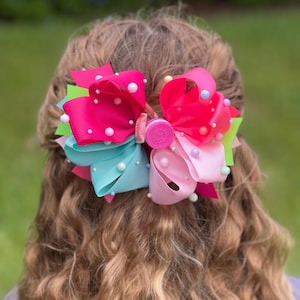 May include: A large, multi-layered hair bow featuring vibrant ribbons in shades of hot pink, turquoise, lime green, and light pink. The bow is embellished with numerous small, round white and pastel-colored beads and has a central circular pink clip.