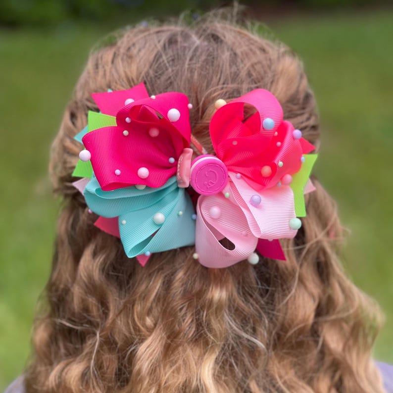 May include: A large, multi-layered hair bow featuring vibrant ribbons in shades of hot pink, turquoise, lime green, and light pink. The bow is embellished with numerous small, round white and pastel-colored beads and has a central circular pink clip.