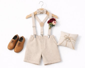 May include: A beige shorts set with suspenders, a bow tie, and brown leather shoes. A small floral arrangement and a ring pillow are also included. The outfit is displayed on a white background.