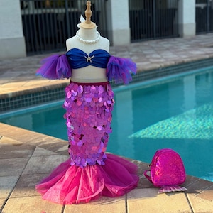 Peut inclure: Un costume de sirène pour enfant, présenté sur un mannequin. Il comprend un haut bleu, une jupe à sequins violets et une queue en tulle rose. Un petit sac à dos rose est également inclus. Le costume est pour enfants.