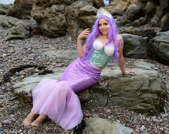 May include: A woman wearing a purple and green mermaid costume with a seashell crown. She is sitting on a rock with a pebbled beach in the background.