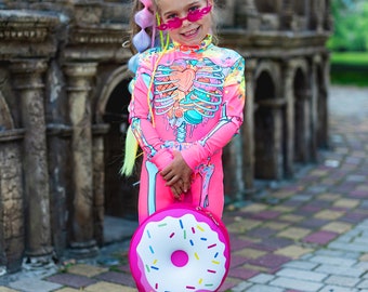 May include: A bright pink long-sleeve jumpsuit featuring a colorful skeleton and candy graphic print, paired with pink flame-shaped sunglasses and a round pink donut-shaped bag with white icing and colorful sprinkles, worn with white sneakers.