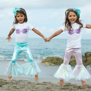 May include: Two girls wearing mermaid costumes, one in blue and one in pink, jump in the air on a sandy beach. They are both smiling and holding hands.